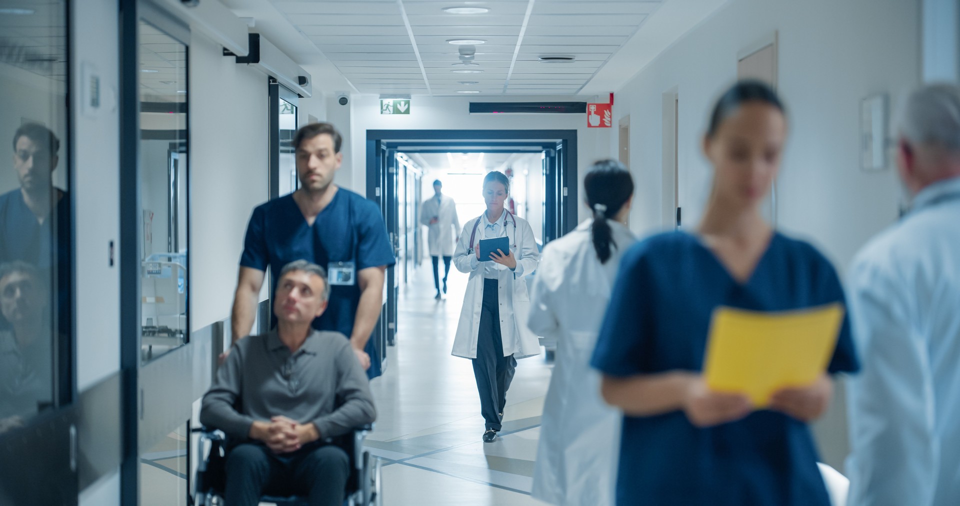 Busy Hospital Hallway Filled with Medical Experts in White Coats and Blue Uniforms. Middle Aged Female Doctor Reviewing a Patient's History on a Tablet Computer as She Heads to the Examination Room